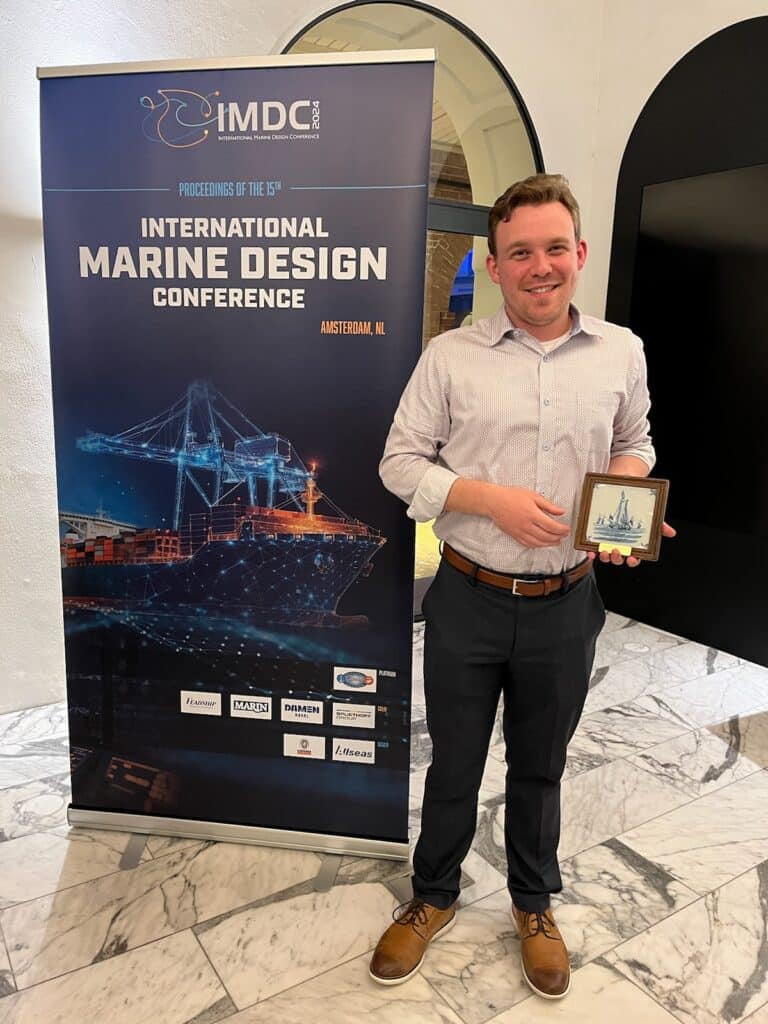 Male student stands in front of International Marine Design Conference sign holding his award.